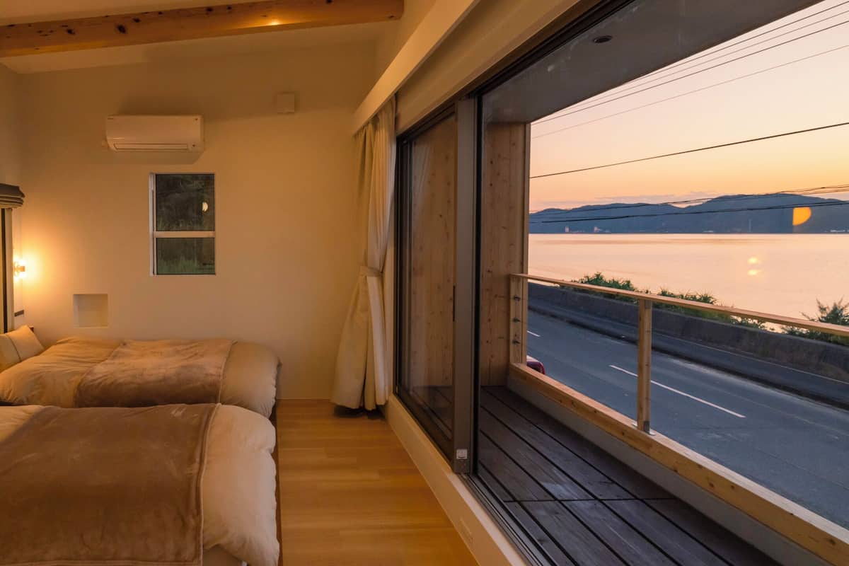 A bedroom with two beds, a large window with a view of a road and mountains, and a wooden ceiling.