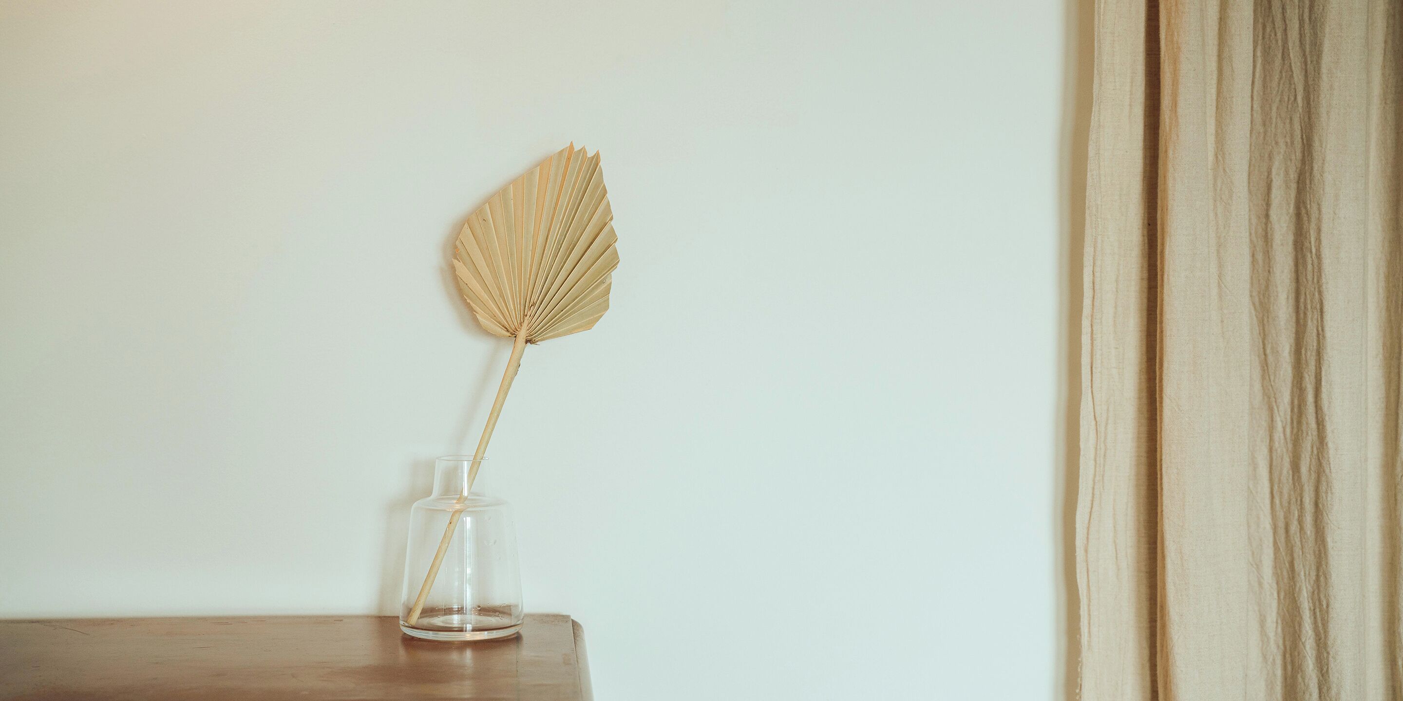 A clear glass vase with a single dried leaf on a wooden surface against a plain wall.