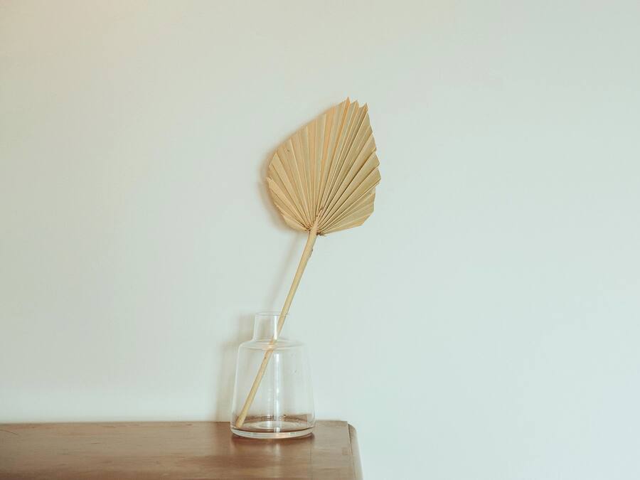 A clear glass vase with a single dried leaf on a wooden surface against a plain wall.