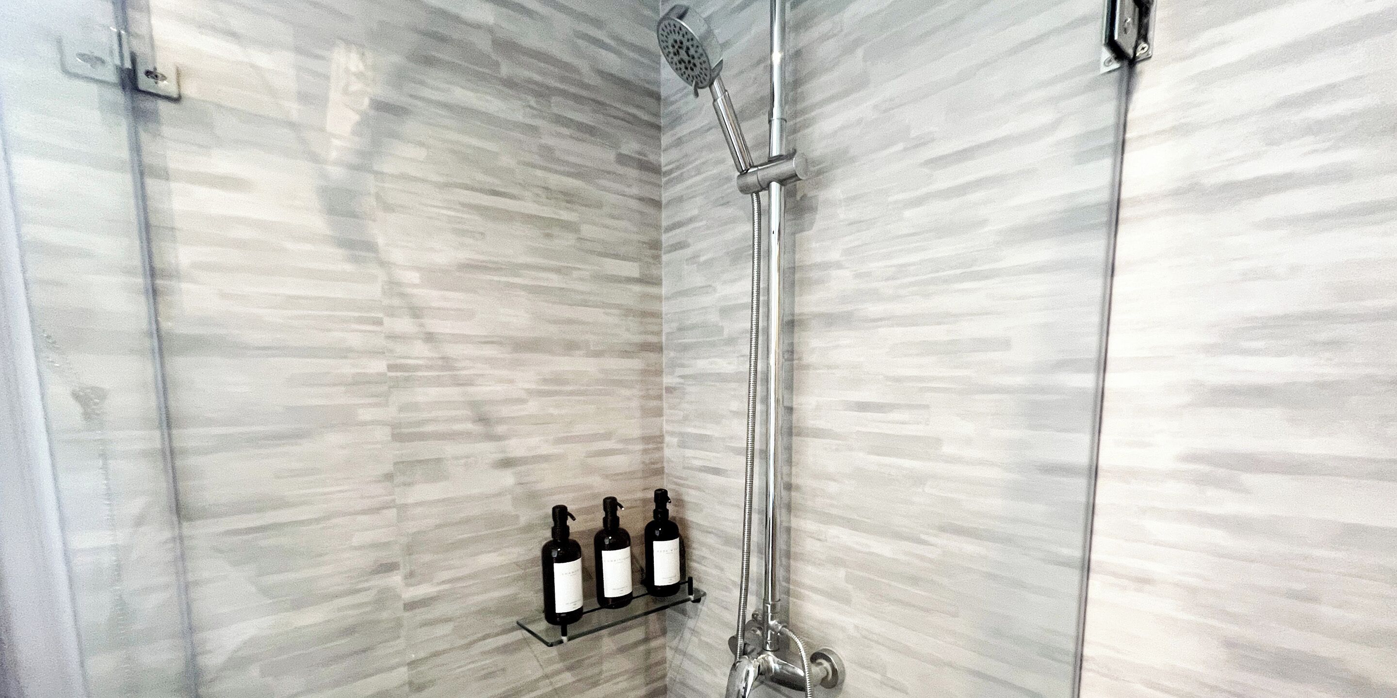A shower with a handheld showerhead and a wall-mounted faucet. There are three bottles on a shelf below the shower.