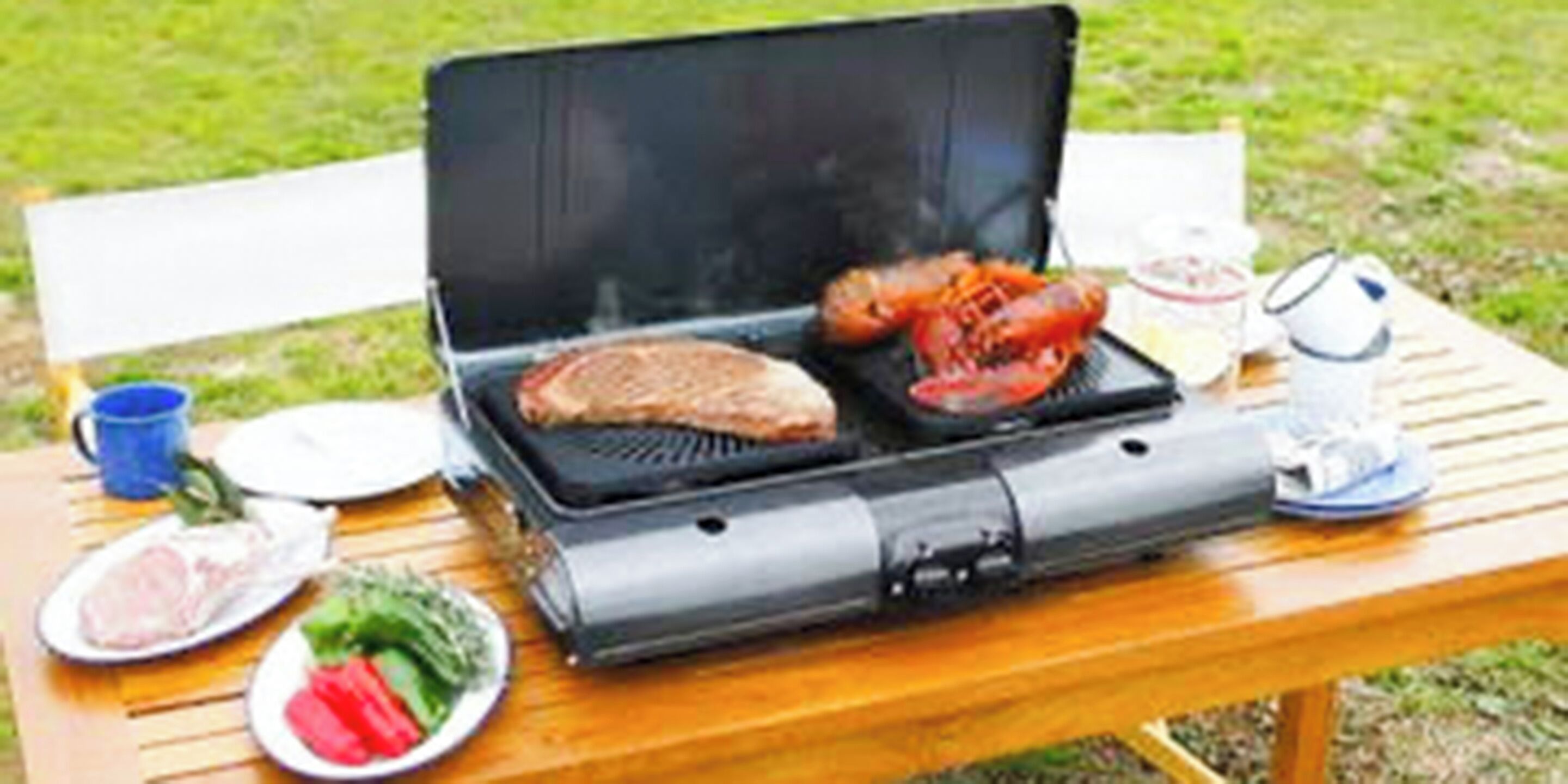 Una parrilla portátil con dos porciones de carne y verduras sobre una mesa de madera al aire libre.