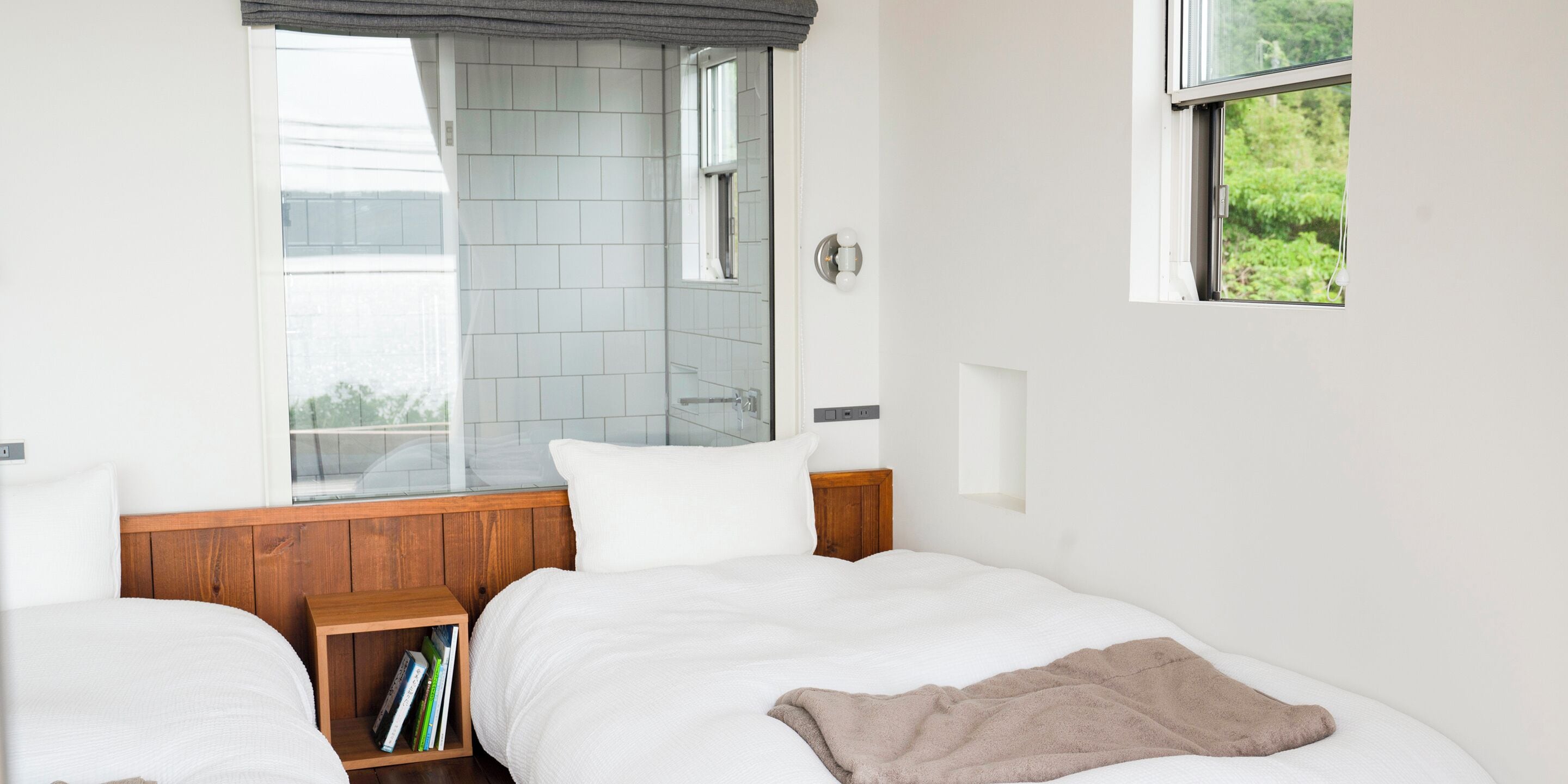 Ein Hotelzimmer mit zwei Betten, einem Holzkopfteil, einem kleinen Nachttisch mit Büchern, einem Fenster mit Blick auf Grünflächen und einem Duschbereich mit Glastür.