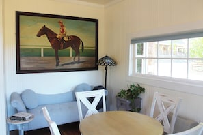A room with a painting of a jockey on a horse, a window with a view, a table, and a sofa. - Historic Cottage at Ridgewood Ranch, Home of Seabiscuit (Willits)
