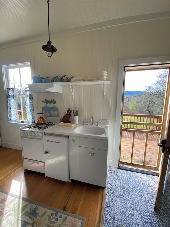 Une cuisine équipée d’une cuisinière à gaz, d’un évier et de meubles blancs. Une fenêtre avec des rideaux offre une vue sur les arbres à l’extérieur.