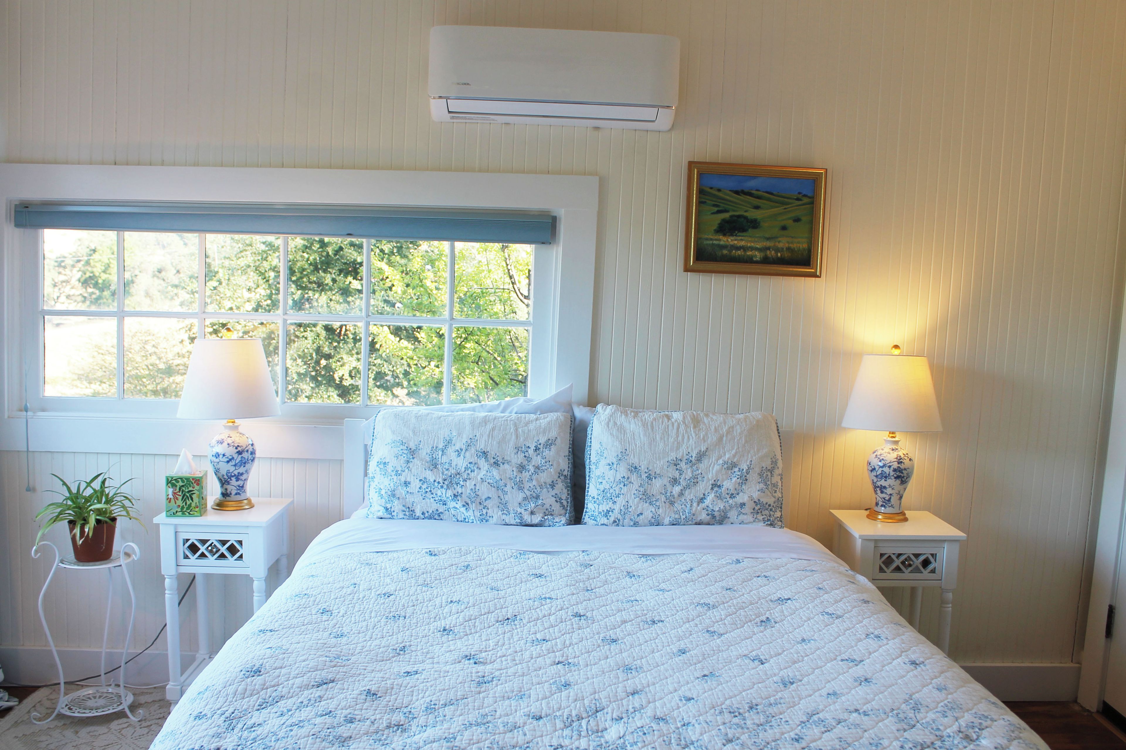 Ein Schlafzimmer mit großem Fenster, zwei Nachttischlampen, einem Wandbild und einem Blumentopf.
