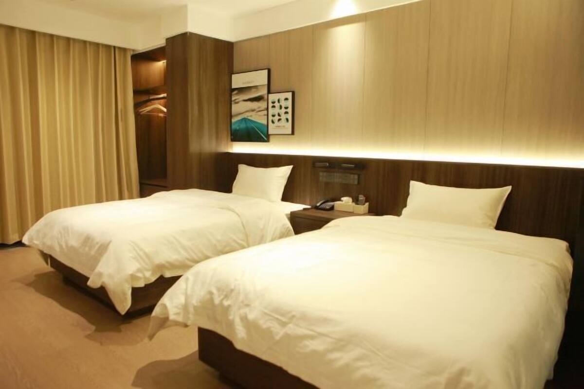 Quarto de hotel com duas camas, cabeceira de madeira, mesa de cabeceira com abajur e um quadro na parede.