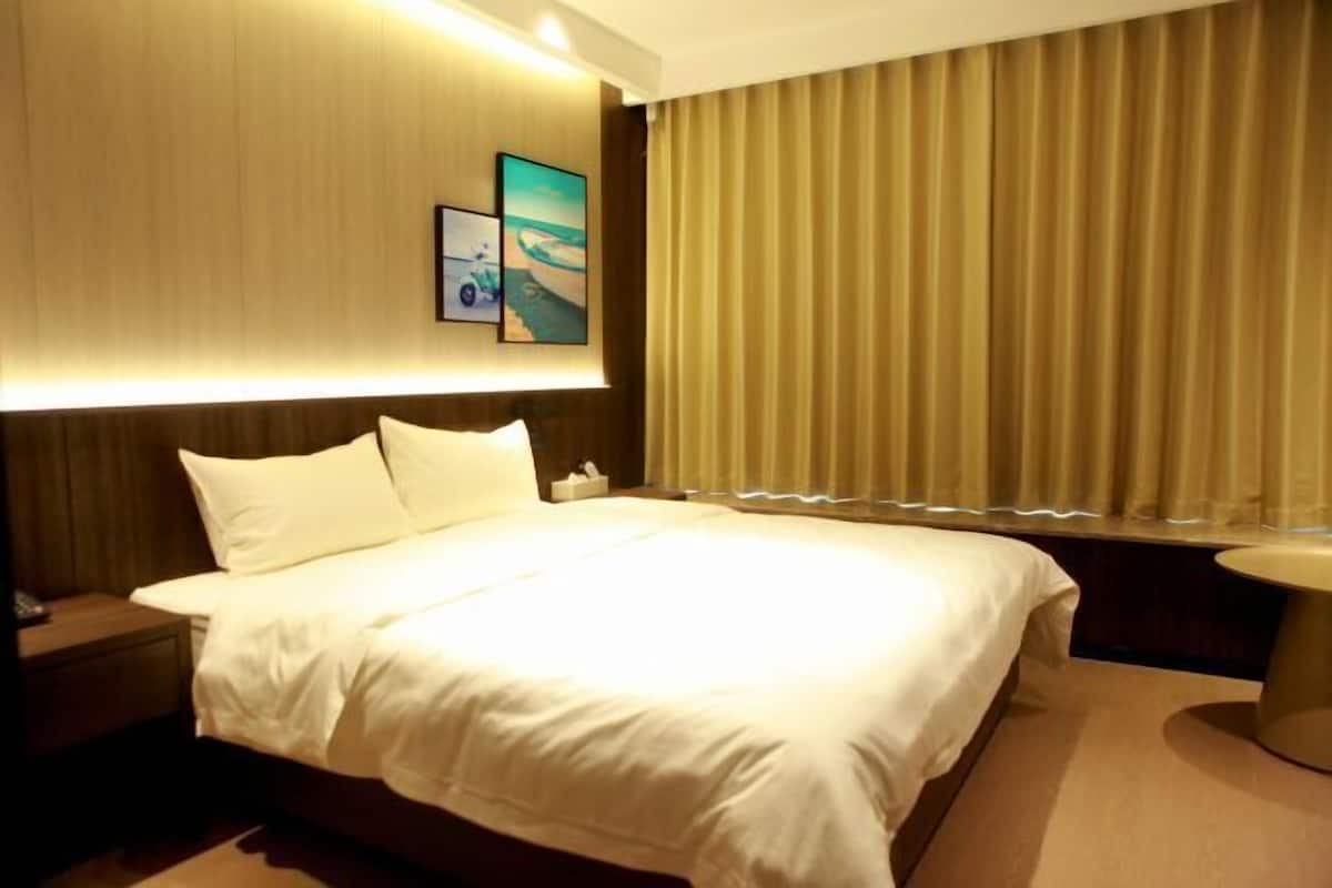 Quarto de hotel com uma cama grande, mesa de cabeceira, uma mesinha redonda e uma janela com cortinas.