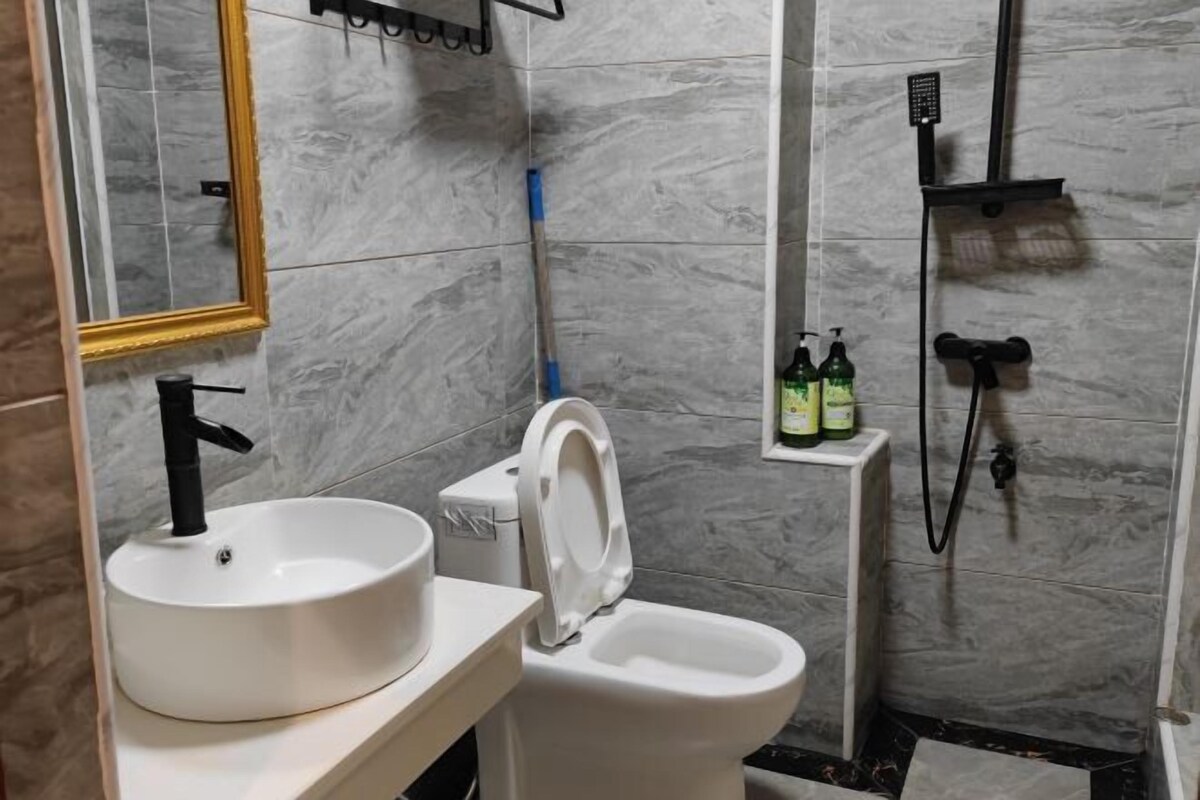 Een badkamer met een wit wastafel, toilet en douche. Er staan flessen shampoo en conditioner aan de muur.