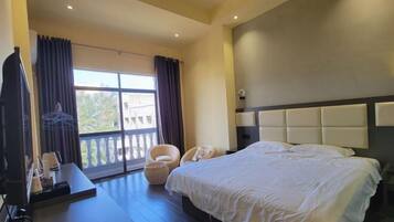 Een hotelkamer met een groot bed, een bureau en een stoel. Er is een balkon met gordijnen en uitzicht op buiten.