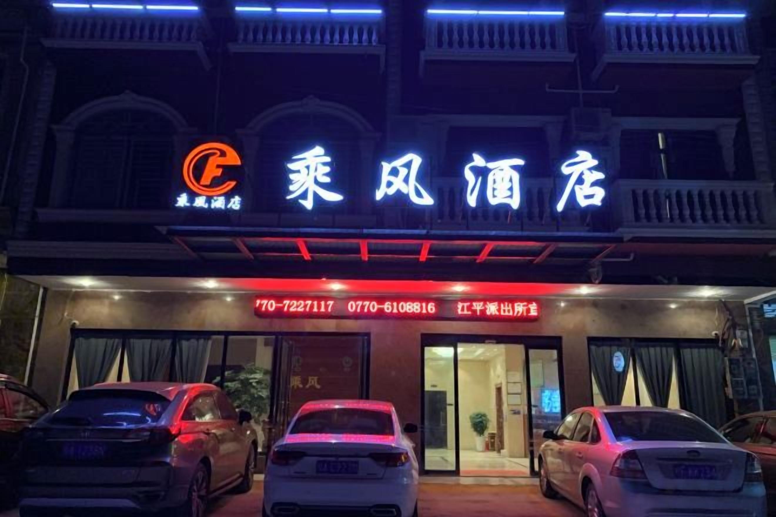 A hotel entrance with a neon sign, a red awning, and a parking area with cars.