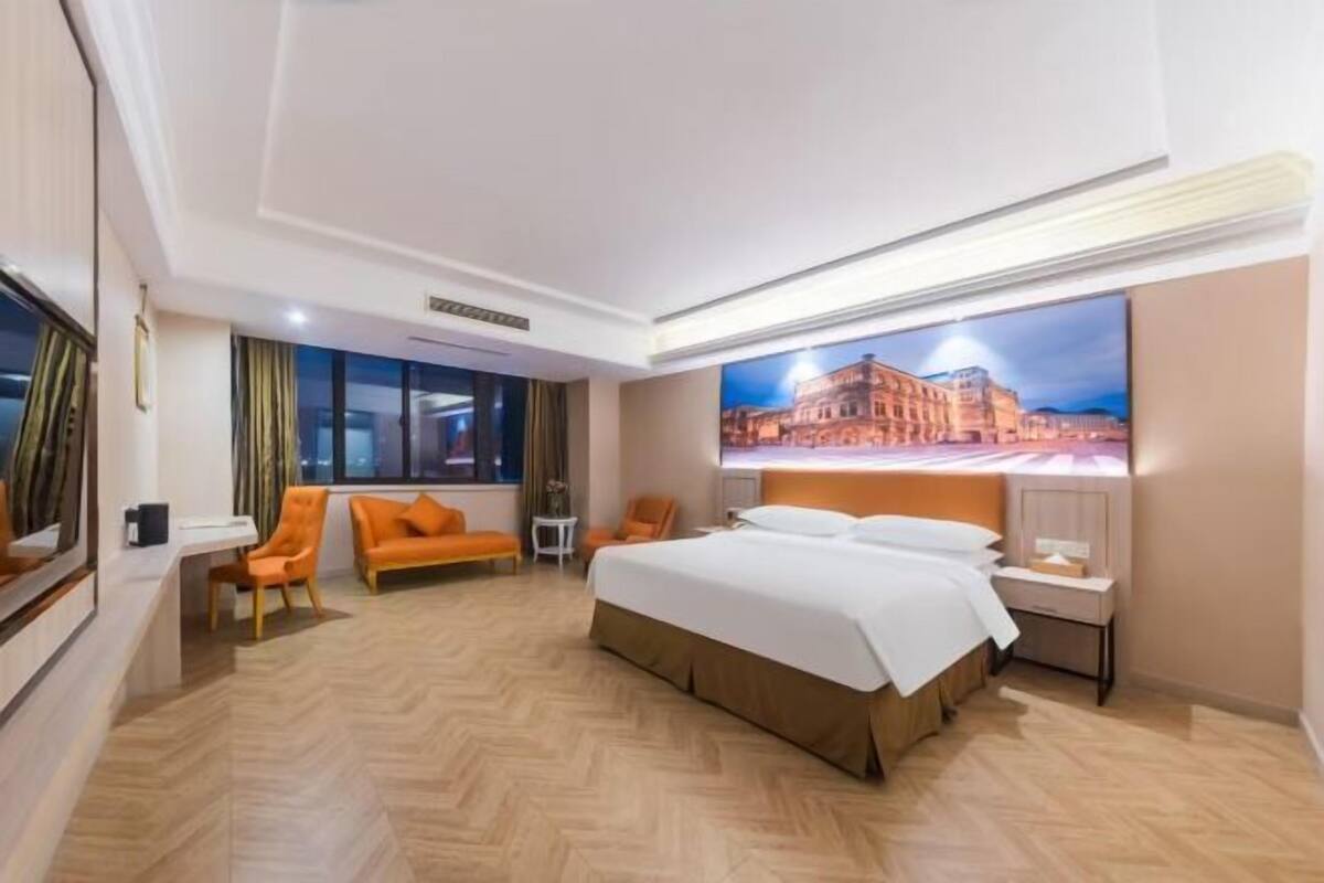A hotel room with a large bed, two chairs, a small table, and a large window.