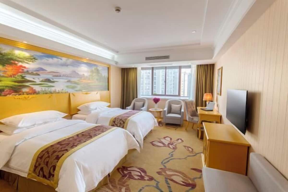 A hotel room with two beds, a TV, a desk, and a window with curtains.