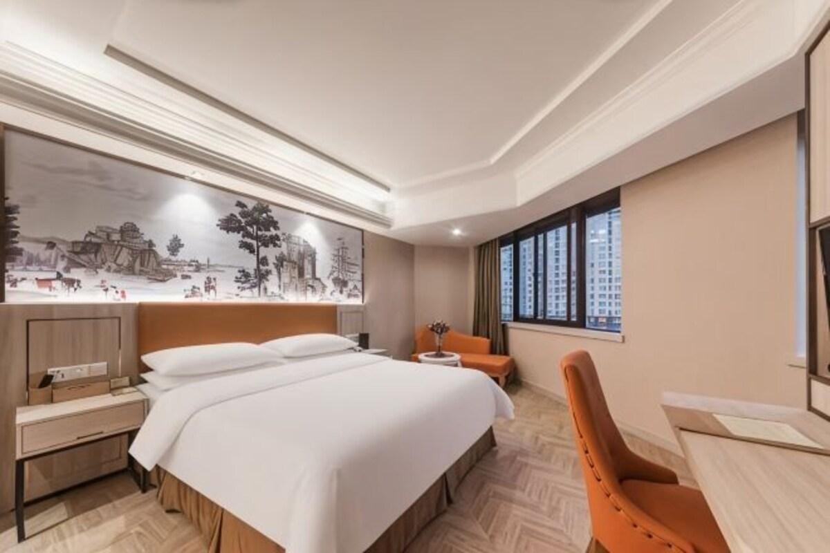A hotel room with a large bed, a desk, a chair, and a window with a view of buildings.