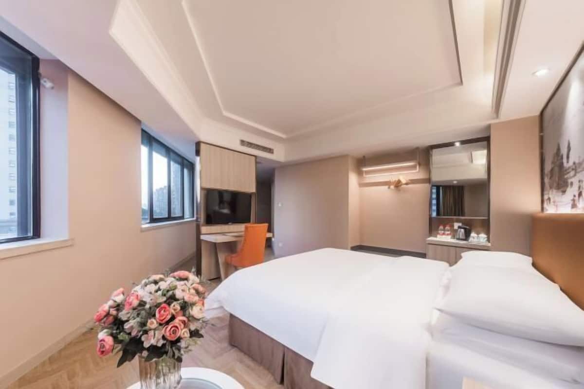 A modern hotel room with a large bed, a desk, and a chair. There is a window providing natural light and a vase of flowers on the desk.