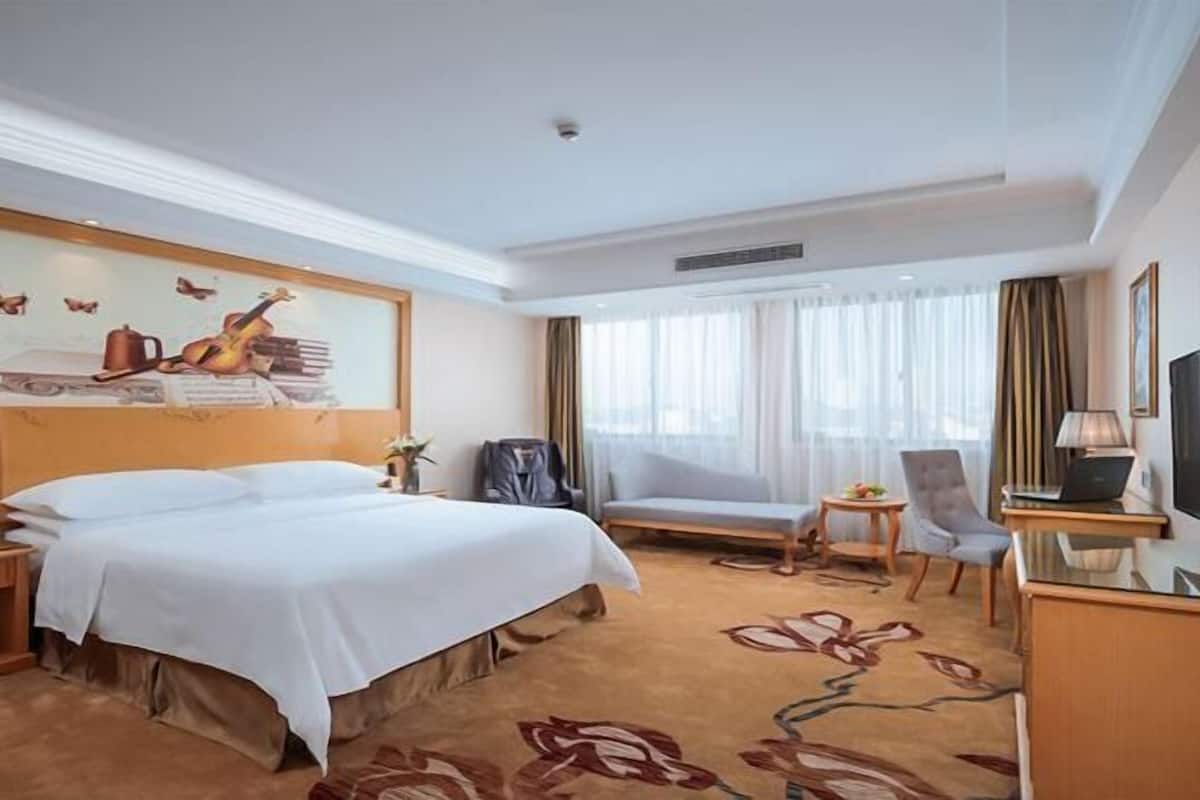 A hotel room with a large bed, a seating area with a chair and a table, a TV, and a window with curtains.