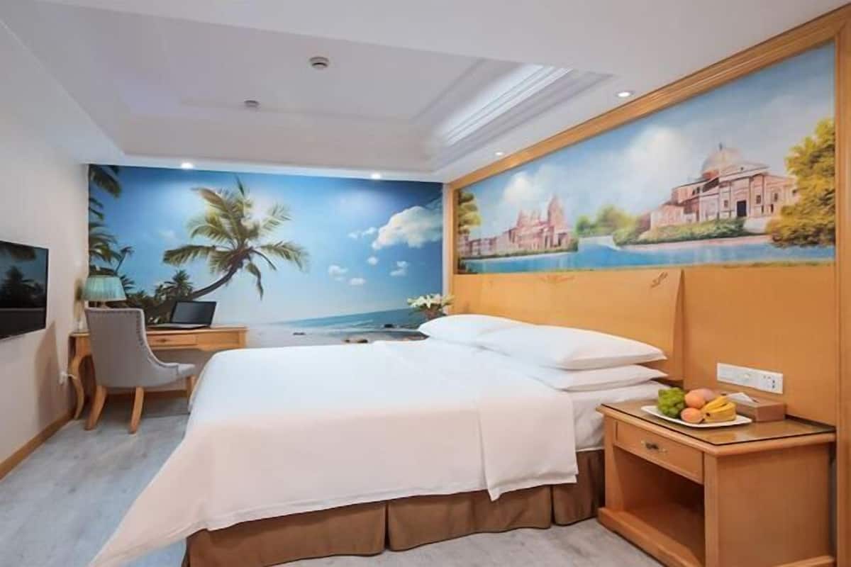 A hotel room with a bed, a desk, a chair, a nightstand with fruit, and a mural of a beach scene.