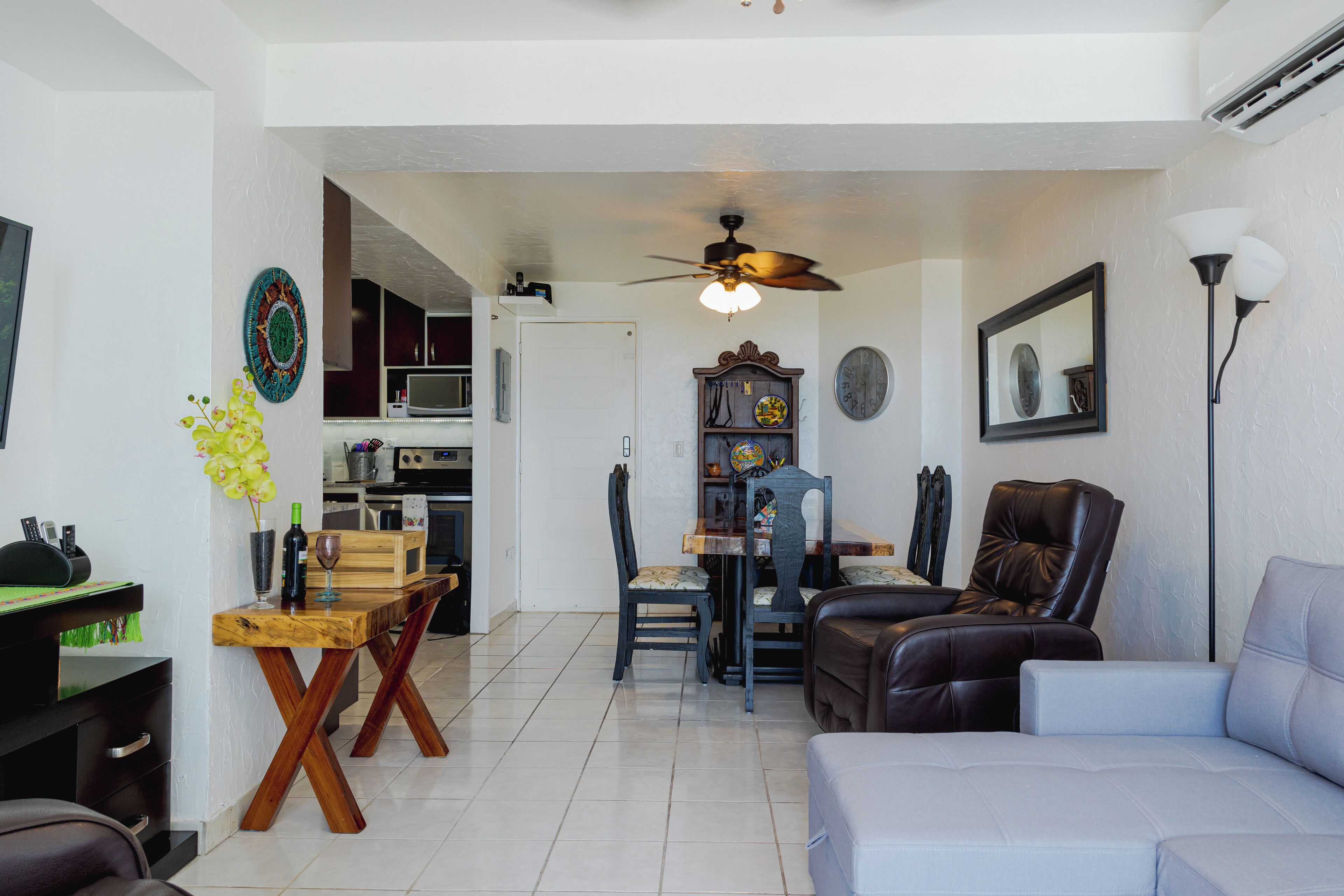 A living room with a sofa, a dining table with chairs, a ceiling fan, and a kitchen area in the background.