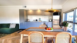 A modern kitchen with grey cabinets, a dining area with a wooden table and wicker chairs, and a view of a building through large windows. - Chic penthouse apartment in heart of Darlinghurst (Darlinghurst)