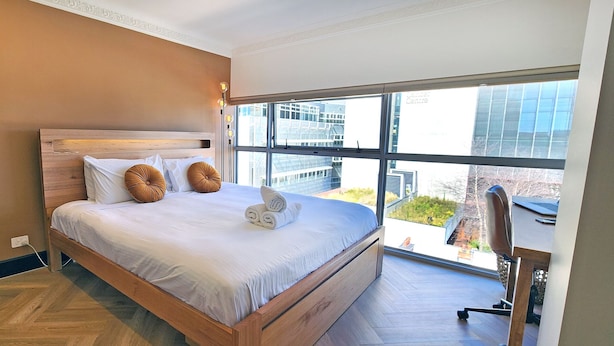 A modern bedroom with a wooden bed, white linens, and a city view through large windows. - Chic penthouse apartment in heart of Darlinghurst (Darlinghurst)