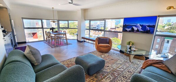 Un salon spacieux avec un grand canapé, un coin repas comprenant une table et des chaises, et une vue sur la ville grâce à de grandes fenêtres.