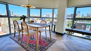 A dining area with a table, chairs, and a bench, large windows with a city view, and a patterned rug. - Chic penthouse apartment in heart of Darlinghurst (Darlinghurst)