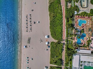 An aerial view of a beach resort with a swimming pool, beach umbrellas, and lounge chairs.