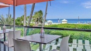 Un balcon avec une table et des chaises, donnant sur une plage bordée de palmiers et offrant une vue sur un ciel dégagé.