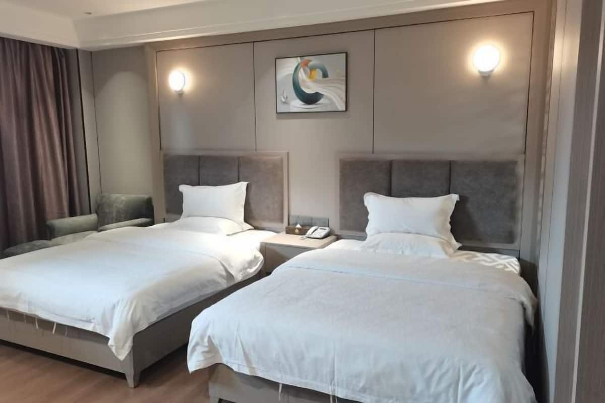 A hotel room with two beds, a bedside table with a phone, a wall-mounted light, and a painting.