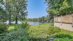 A lakeside view with a wooden deck, lush greenery, and a clear sky. - Picturesque Phoenix Vacation Home on Oswego River! (Phoenix)