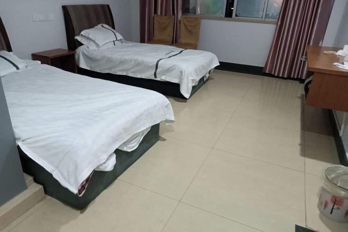 A hotel room with two single beds, a wooden desk, a chair, a window with curtains, and a trash bin.