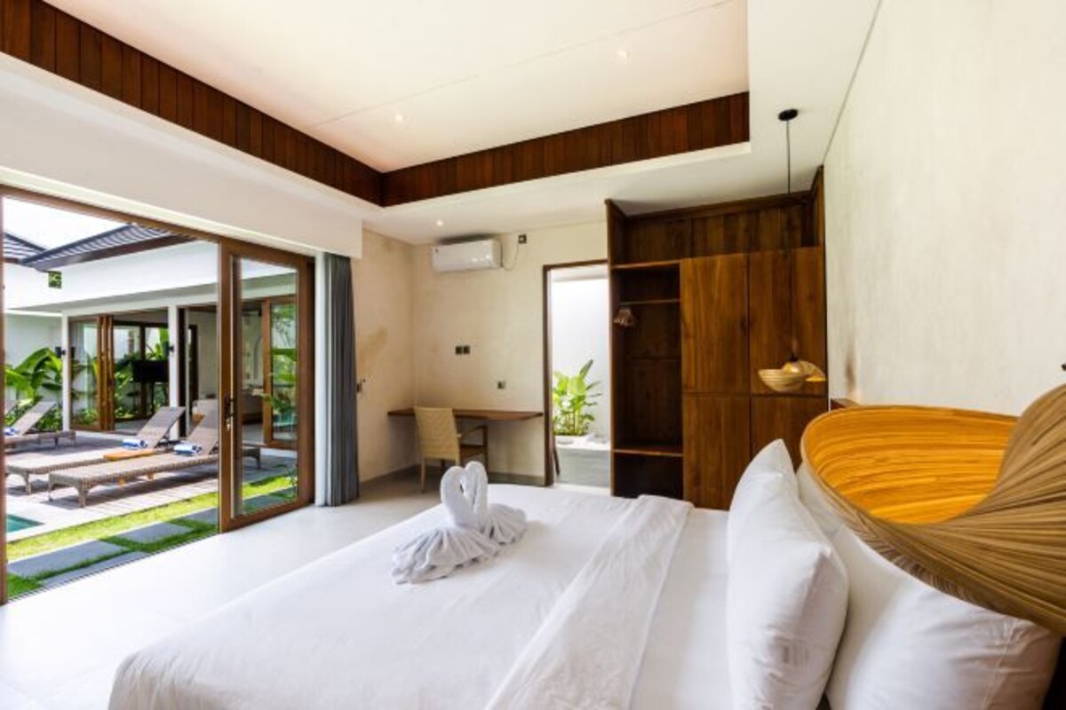 Une chambre d’hôtel moderne dotée d’un grand lit, d’une tête de lit en bois, d’un bureau et offrant une vue sur une piscine et un jardin.