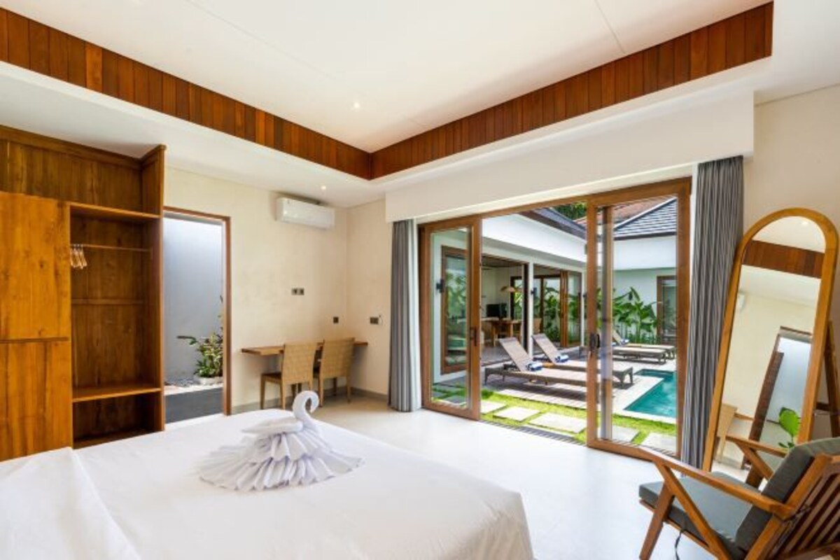 A modern hotel room with a large bed, a wooden wardrobe, a desk, and a view of a pool and garden through glass doors.