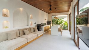 A modern living room with a wooden ceiling, a sofa, a dining area with chairs, and a kitchen area with a refrigerator and coffee machine. - J4 Villas Sanur (Denpasar)