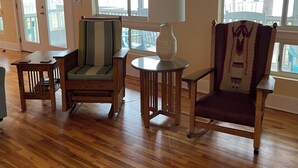 A room with wooden flooring, a chair with a patterned cushion, a small table with a lamp, and a clock on the wall.