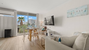 Un salon avec balcon, un téléviseur fixé au mur et une vue sur des palmiers et des immeubles.
