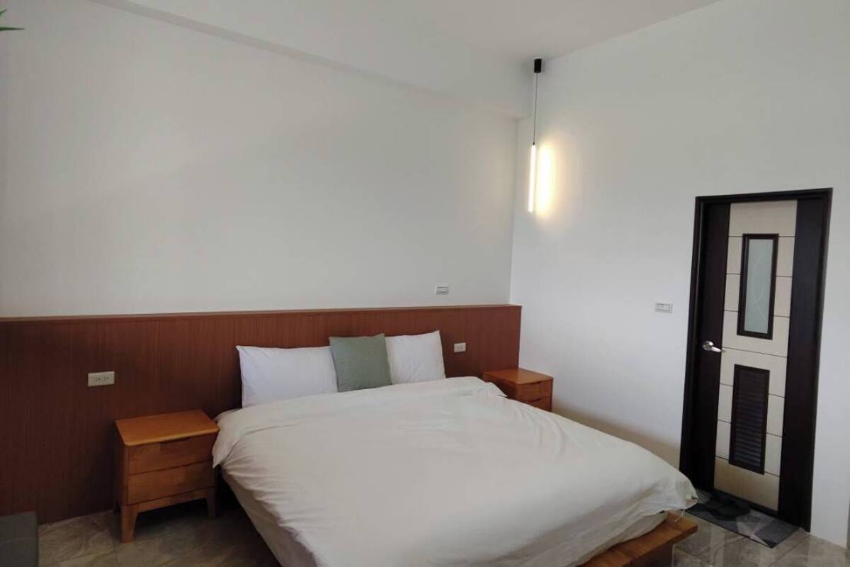 A modern bedroom with a wooden headboard, a single bed, a bedside table, a nightstand, a wall-mounted light, and a door with a mirror.
