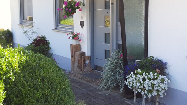 Ein Haus mit weißer Fassade, einer grauen Tür und einem Fenster mit einem Blumenkasten. Draußen stehen Blumentöpfe und ein Holzschemel.