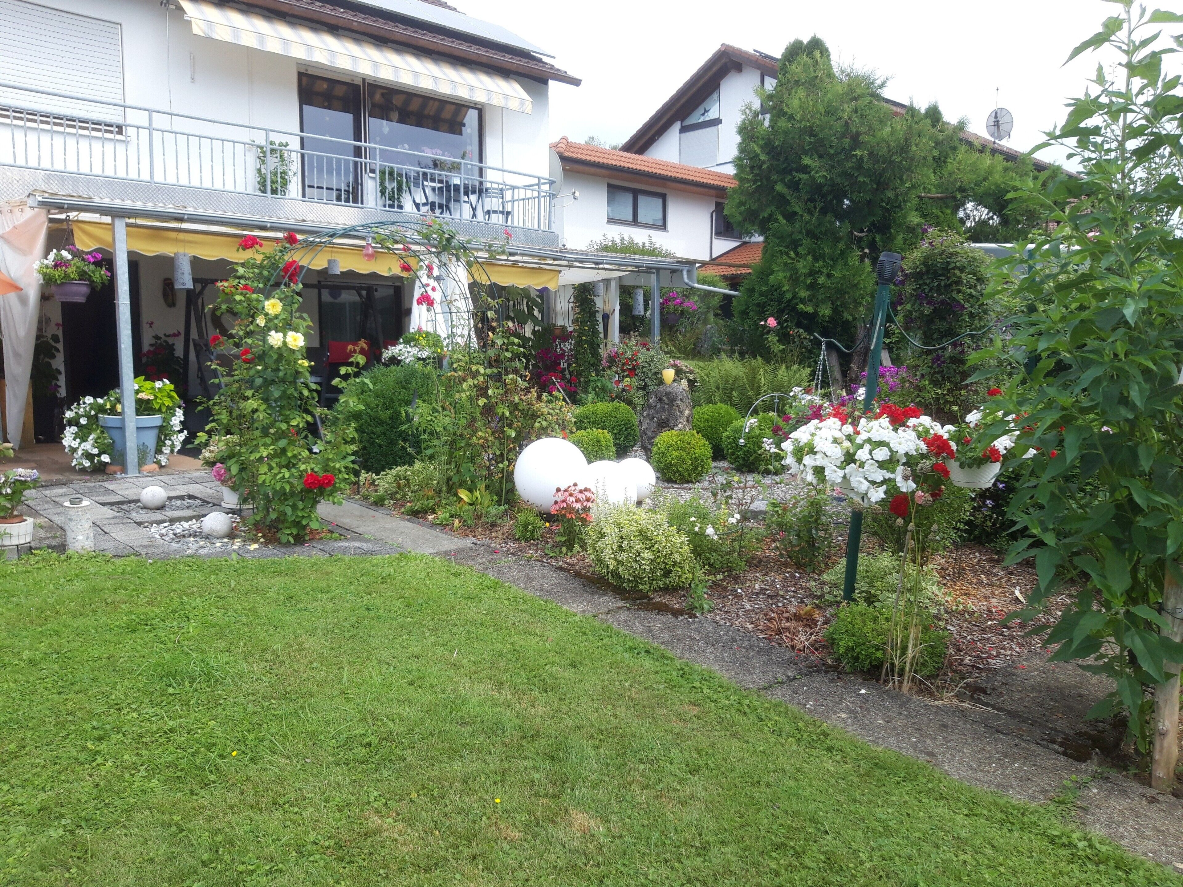 Une maison à deux étages avec un balcon, un jardin agrémenté de diverses plantes et fleurs, et un chemin menant à l’entrée.