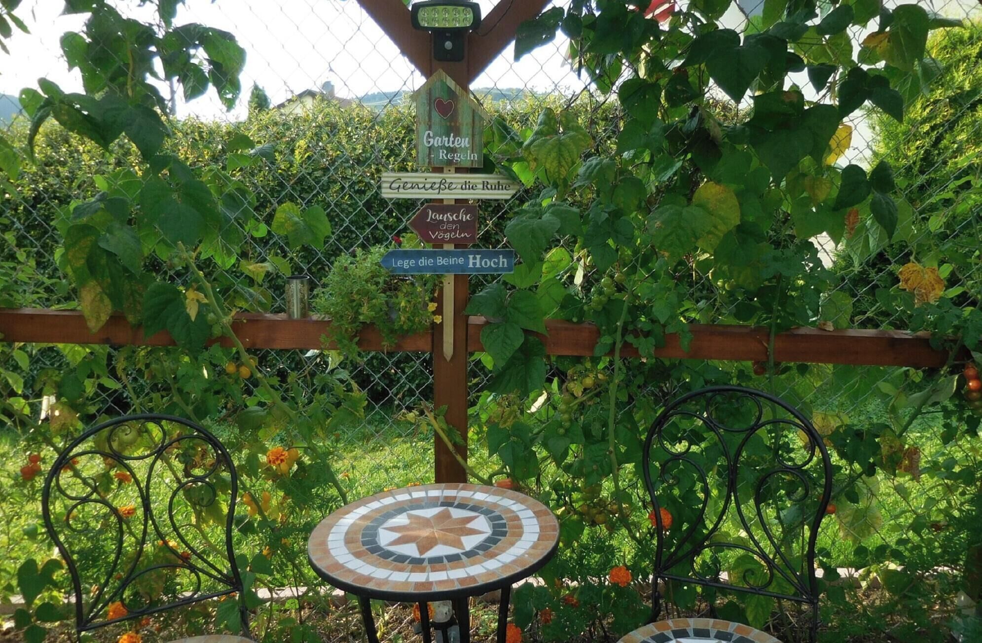 A garden with a wooden fence, a metal table with a mosaic top, and various signs in a foreign language.