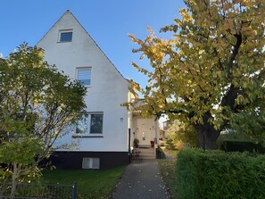 Ein weißes, zweistöckiges Haus mit einer sichtbaren Haustür, einem kleinen Fenster und einem gepflasterten Weg zum Eingang.