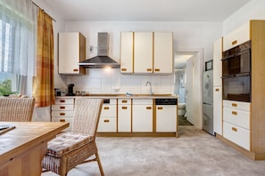 A modern kitchen with white cabinetry, a built-in stove, and a dining area with a wicker chair.