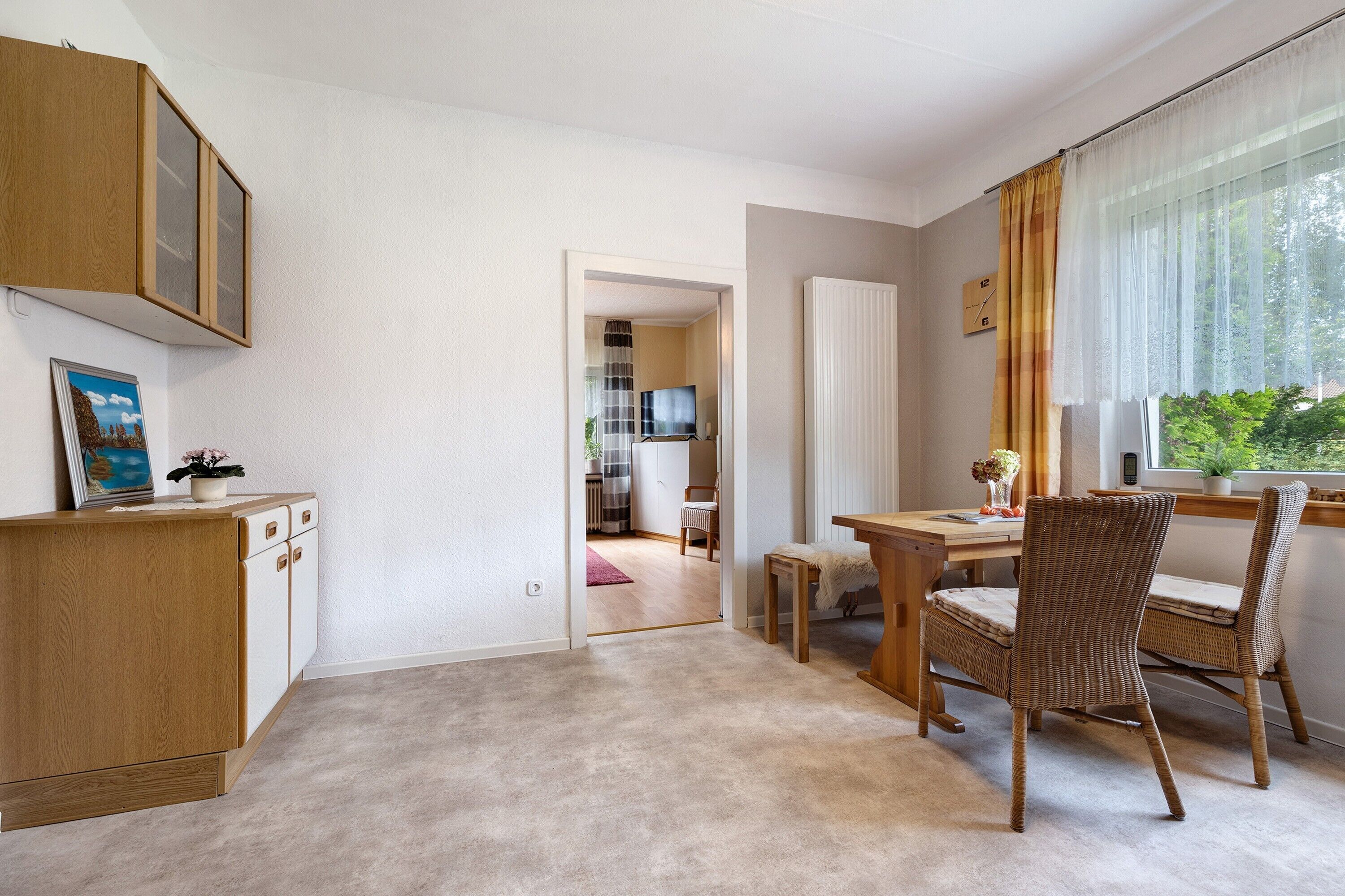 Ein Zimmer mit Holzschrank, Esstisch mit Stühlen, Fernseher im Hintergrund und einem Fenster mit Vorhängen.