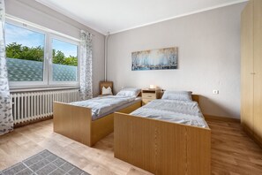 Et soveværelse med to senge i træ, en radiator, et vindue med gardiner, et billede på væggen og et lille natbord.