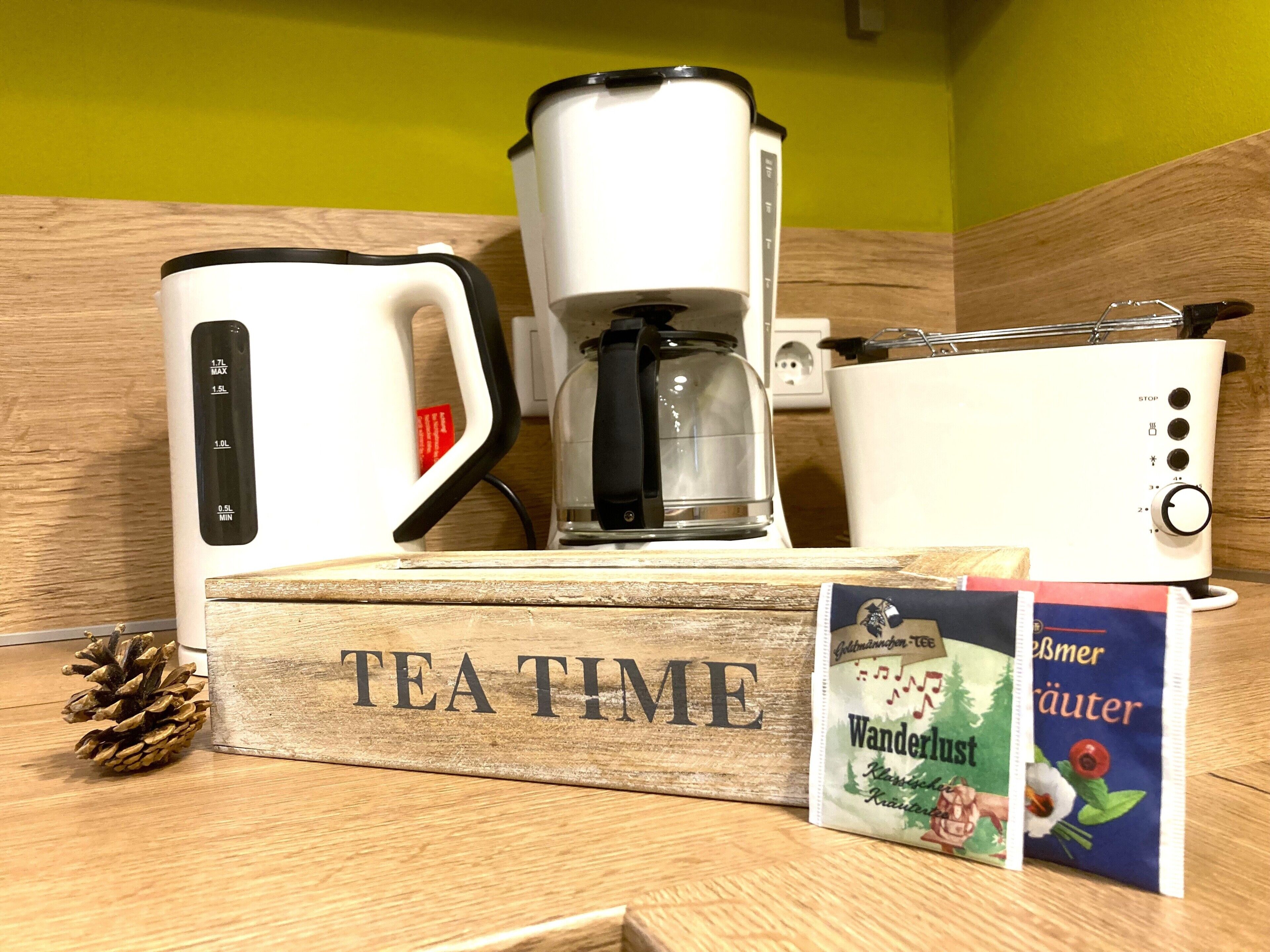 Eine Kaffeemaschine, ein Wasserkocher und ein Teebeutel auf einer Holztischplatte, mit der Aufschrift 'TEA TIME'.