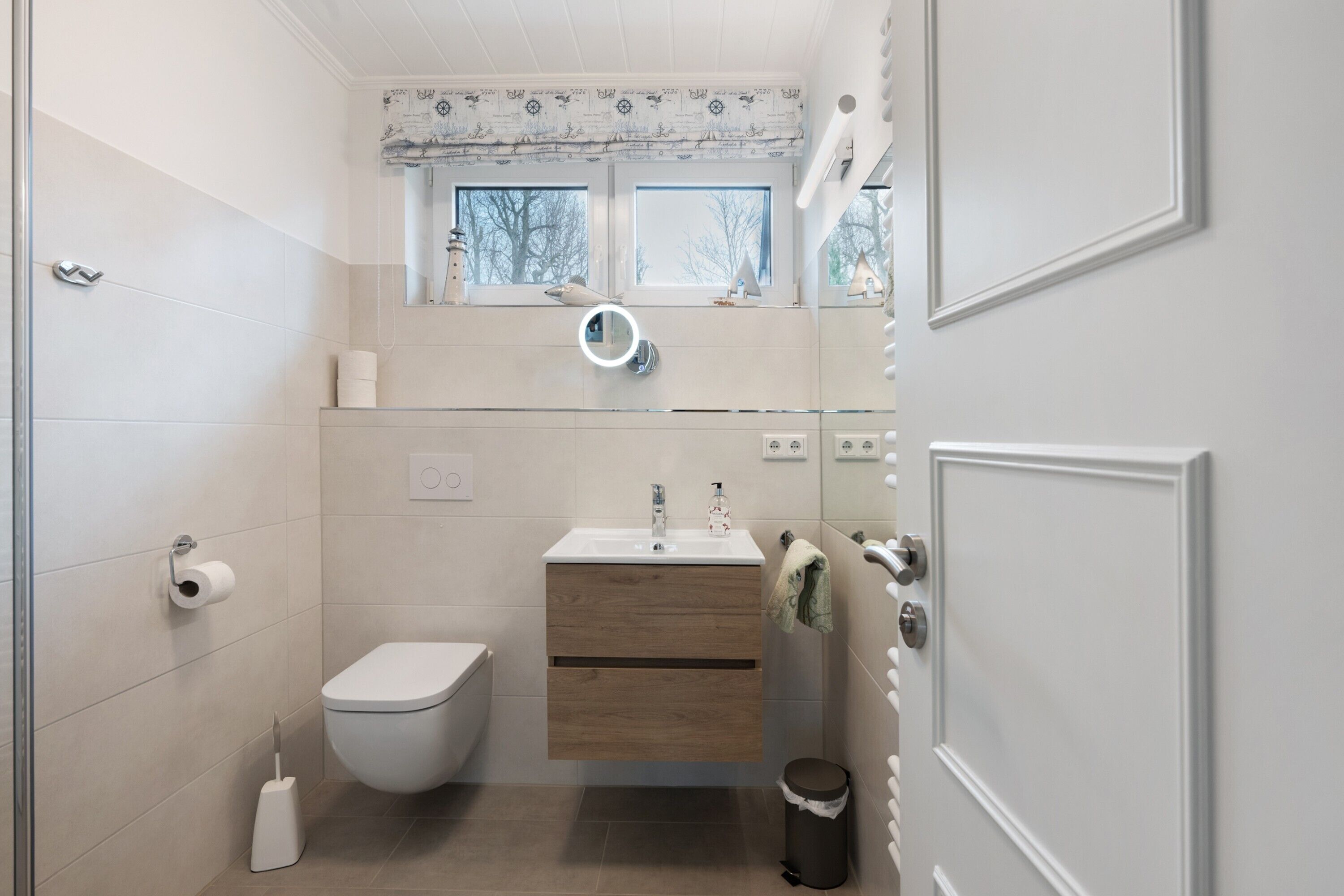 Et badeværelse med toilet, vask, underskab og et vindue med udsigt til træer.