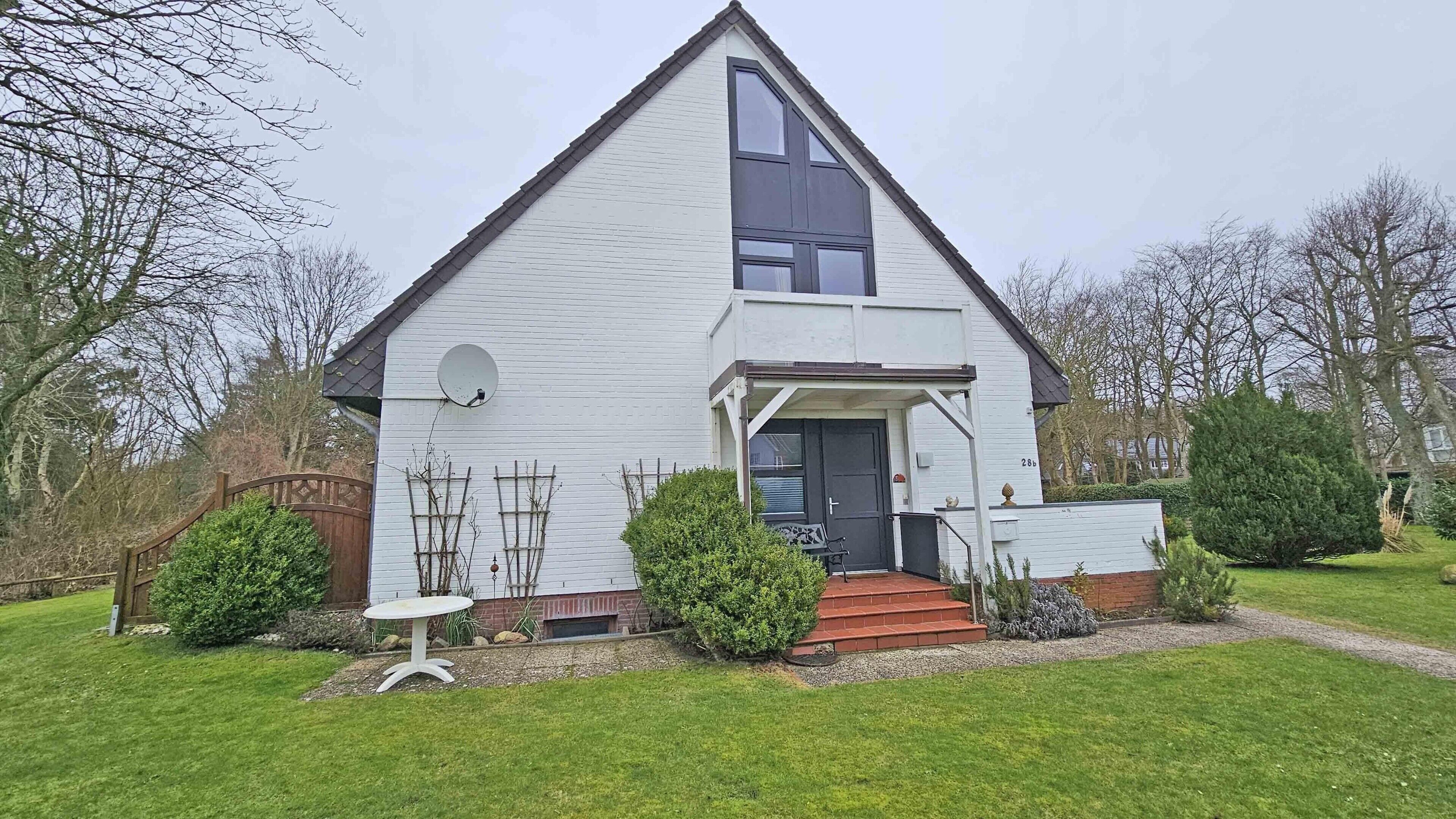 Une maison blanche à deux étages, avec une porte noire, une antenne parabolique et un petit jardin agrémenté d’une table et de chaises.