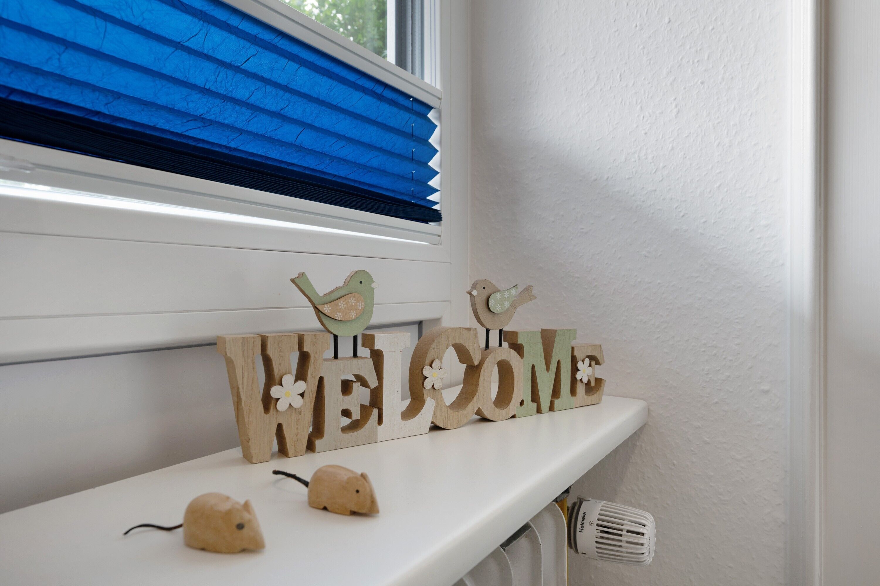Une étagère blanche ornée d’une pancarte « BIENVENUE », de deux figurines d’oiseaux en bois et de deux petites figurines d’animaux.