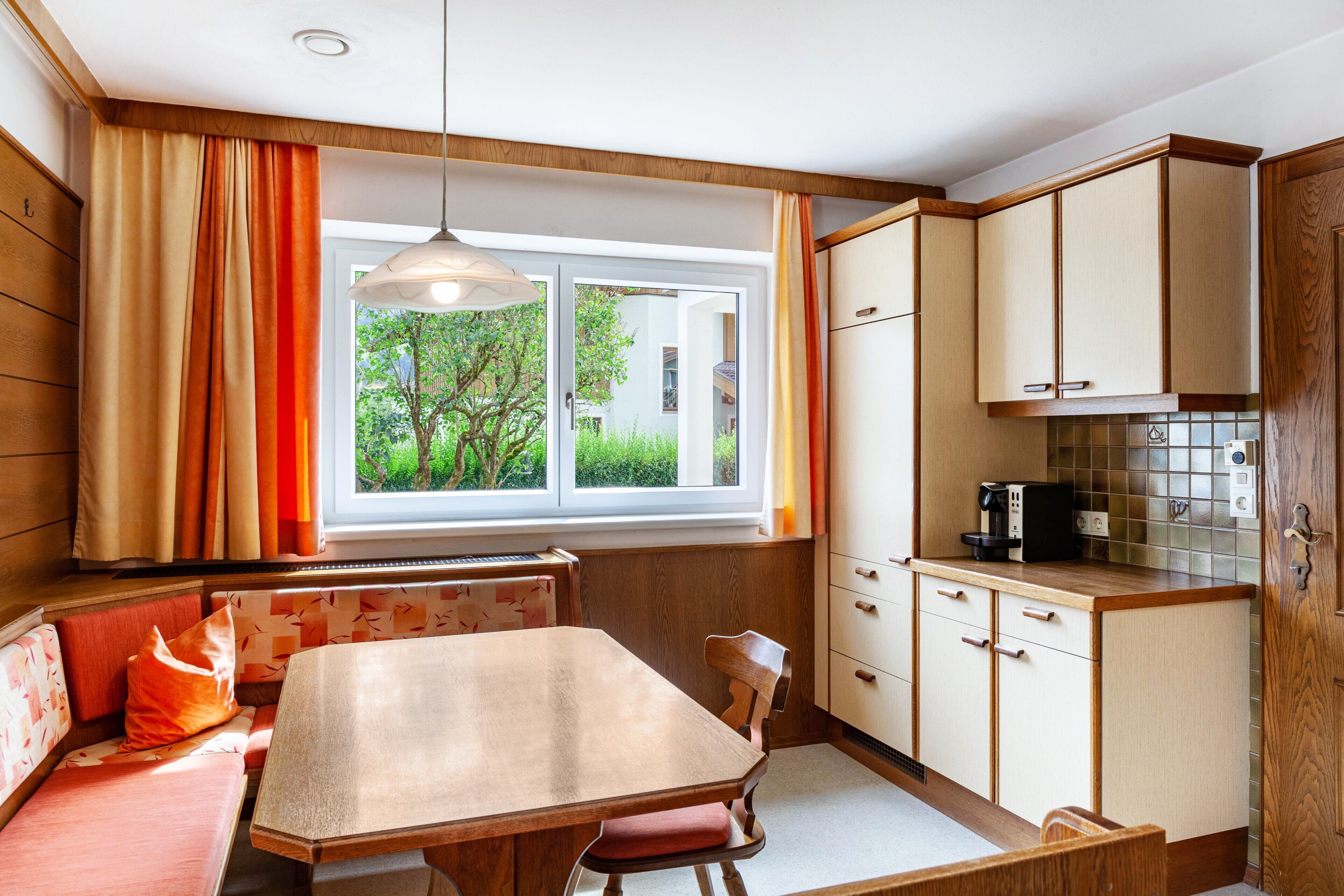Eine Küche mit Esstisch, Holzschränken, einem Fenster mit Vorhängen und Blick ins Grüne.
