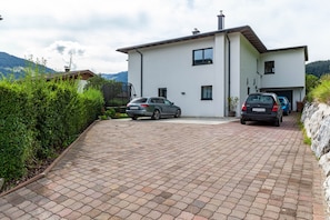Una casa moderna de dos pisos con entrada pavimentada, coches estacionados y vistas a la vegetación y colinas circundantes.