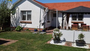 Een gelijkvloers huis met een rood dak, witte buitenmuren en een kleine voortuin met potplanten en een terras.